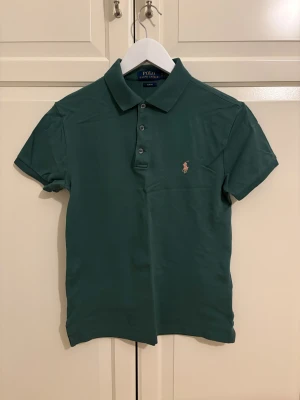 Grön slim fit piké från Polo Ralph Lauren - Snygg grön pikétröja från Polo Ralph Lauren i slim fit-modell. Klassisk krage, tre knappar framtill och den ikoniska broderade loggan på bröstet. Tillverkad i mjuk bomull som är skön mot huden. Perfekt för dig som gillar stilrena och sportiga plagg.