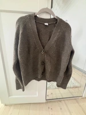 Brun stickad kofta med knappar - Mysig brun stickad kofta från Lindex med tre stora knappar framtill och v-ringad hals. Koftan har långärmade ärmar och ribbade muddar vid ärmslut och nederkant. Perfekt för att hålla sig varm under kyliga dagar.