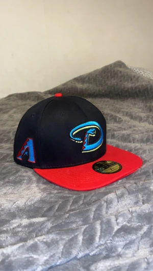 Arizona Diamondbacks keps New Era - Snygg Arizona Diamondbacks keps från New Era 59FIFTY med svart krona och röd skärm. Broderad logga i blått, gult och rött framtill samt MLB-märke bak. Klassisk flatbrim och röd knapp på toppen. Perfekt för dig som gillar baseball och streetwear.