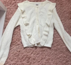 Vit kofta med volanger och knappar - Supersöt vit kofta med volangdetaljer längs axlar och framstycke. Stickad struktur och knappar framtill ger en extra touch. Perfekt att styla med jeans eller kjol för en trendig look. Långärmad och ribbad vid muddar och nederkant.