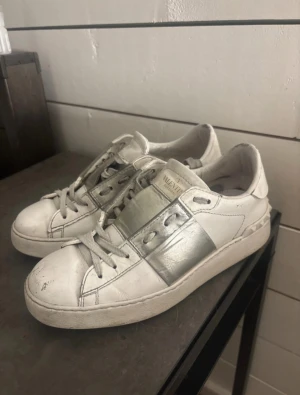 Valentino Garavani vita och silver sneakers - Snygga sneakers från Valentino Garavani i vitt skinn med en bred metallic-silver detalj över sidan. Klassisk rund tå, platt sula och snörning framtill. Ikoniska nitar baktill på sulan ger extra edge. Perfekt för dig som gillar lyxiga och trendiga skor.