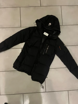 Svart Sail Racing dunjacka  - Svart dunjacka från Sail Racing. Jackan har huva, flera dragkedjor och snygga logodetaljer på bröstet och ärmen. Perfekt för kalla dagar med sin puffiga design och sportiga look. Passar dig som gillar stil och funktion. Vanliga kedjan är borttappad och därför ersatt med en liten ring men jackan går att öppna och stänga som vanligt ändå. Finns ett litet hål som visas bland bilderna. 