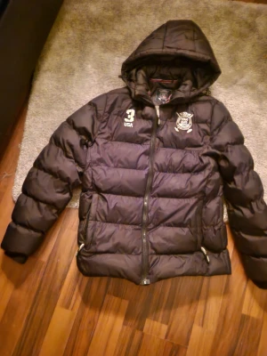 Svart pufferjacka från Vinson Polo Club - Svart pufferjacka med huva från Vinson Polo Club. Jackan har broderade detaljer på bröstet, dragkedja framtill och två sidofickor med dragkedja. Perfekt för kalla dagar och har en sportig look med USA-tryck och klubbmärke. ORDINARIE PRIS 550KR