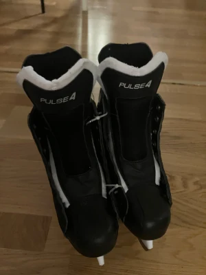 Svarta Pulse4 skridskor strl 37 - Säljer ett par svarta och vita skridskor från Pulse4 i storlek 37. De har en robust design med snörning och vadderad plös i vitt syntetmaterial. Yttersulan är vit och skridskorna har detaljer i silver. Perfekt för dig som vill glida fram på isen med stil. Men tyvärr utan snören. 