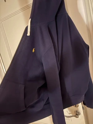 Marinblå hoodie från Polo Ralph Lauren - Snygg marinblå hoodie från Polo Ralph Lauren med gul broderad logga på bröstet. Tröjan har huva med vita snören, dragkedja och en stor ficka framtill. Perfekt för chill dagar och enkel att matcha med jeans eller joggers.