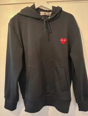 Svart hoodie Comme des Garçons Play - Svart hoodie från Comme des Garçons Play med det ikoniska röda hjärtat med ögon broderat på bröstet. Tröjan har huva med snörning, stor magficka och långa ärmar. Mjuk som är skön att ha på sig. Perfekt för dig som gillar streetwear och stilrena detaljer.
