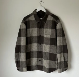 Rutig kort rock från Uniqlo, storlek L - Kort rock från Uniqlo i storlek L med stora rutiga block i grått och svart. Skjortan har klassisk krage, knäppning framtill och en bröstficka. Tillverkad i en blandning av polyester och viskos, perfekt för lager-på-lager-stil. 5K5