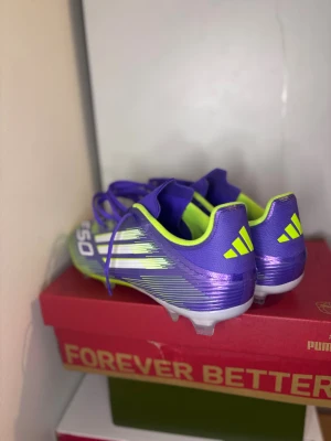 Adidas F50 lila fotbollsskor - Säljer ett par Adidas F50 elite  fotbollsskor i en riktigt fet lila färg med neon-gula detaljer och vita ränder. Skorna har snörning och en platt sula med dobbar för gräsplan. Materialet är syntet och designen är sportig med coola grafiska linjer. Skorna har aldrig använts! Super bra skick.