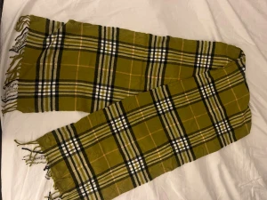 Burberry halsduk - Snygg rutig halsduk från Burberrys i 100% lammull. Färgerna är olivgrön, svart, vit och beige med klassiskt rutmönster och fransar i kanterna. Perfekt accessoar för att lyfta din outfit med en tidlös brittisk vibe.