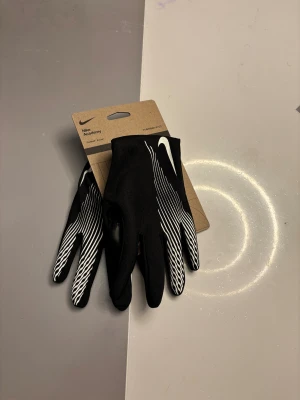 Nike Academy svarta fotbollshandskar - Svarta Nike Academy handskar med vita ränder och Nike-logga på ovansidan. Slimmad passform med Therma-FIT material som håller händerna varma. Snyggt randigt mönster på fingrarna och greppvänlig insida, perfekta för fotbollsträning under kyliga dagar.
