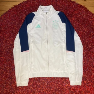 Vit Ajax vindjacka  - Snygg vit vindjacka från Adidas i storlek XS med marinblå detaljer på axlarna och mintgröna ränder på ärmarna. Jackan har dragkedja framtill, elastiska muddar och en cool logga på bröstet. Perfekt för dig som gillar sportig stil och streetwear.