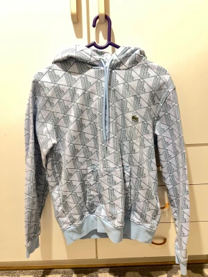 Ljusblå mönstrad hoodie från Lacoste - Säljer en ljusblå hoodie från Lacoste med geometriskt mönster i grått. Klassisk passform, huva med snörning och känguruficka framtill. Ikoniska krokodilloggan på bröstet. Perfekt för dig som vill ha en snygg och bekväm streetstyle.