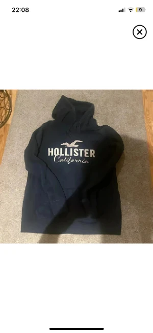 Mörkblå Hollister hoodie med tryck - Snygg mörkblå hoodie från Hollister med stort vitt tryck på bröstet och klassisk logga. Tröjan har huva med snörning och en skön, mjuk insida. Perfekt för dig som gillar en avslappnad och stilren look. Strl L men liten så passar fint på s/m lite oversized❤️