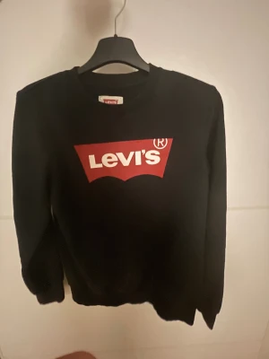 Svart Levi's sweatshirt med röd logga - Svart sweatshirt från Levi's med klassisk röd logga och vit text framtill. Tröjan har rund hals, långa ärmar och ribbade muddar vid ärmslut och nederkant. Perfekt för dig som gillar streetwear och vill ha en enkel men snygg look.