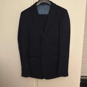 Mörkblå kostym Dressmann Slim Fit - Säljer en stilren mörkblå kostym från Dressmann i slim fit-modell. Klassisk kavaj med två fickor fram och smal passform. Perfekt för dig som vill ha en clean och modern look. Storlek 44 Regular på kavajen och 30-32 på byxorna. Använd 1 gång å det var på balen 