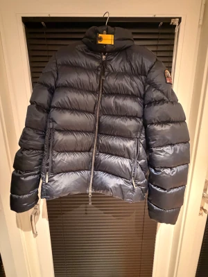 Mörkblå Parajumpers pufferjacka S - Säljer en mörkblå pufferjacka från Parajumpers, modell Sheen. Jackan har en hög krage, tvåvägsdragkedja framtill och sidofickor med dragkedja. Materialet är glansigt och syntetiskt, perfekt för kalla dagar. Klassisk patch på ärmen och orange detalj vid halsen.