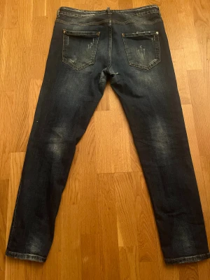 Dsquared2 mörkblå jeans med slitningar - Snygga mörkblå jeans från Dsquared2 med coola slitningar och färgstänk på framsidan. Klassisk femficksmodell med raka ben och låg midja. Jeansen har en vintage tvätt och detaljer som ger en edgy vibe. Perfekt för dig som gillar streetstyle och vill sticka ut. Jeansen är helt äkta.