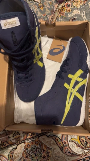 Mörkblå och guldiga  Asics ex eo brotningsskor  - Snygga ex eo från Asics i mörkblått med limegula detaljer på sidorna. Skorna har snörning och en klassisk rund tå. Materialet är syntet med inslag av mocka för en cool look. Perfekt för dig som gillar sportig stil och vill sticka ut med färg.