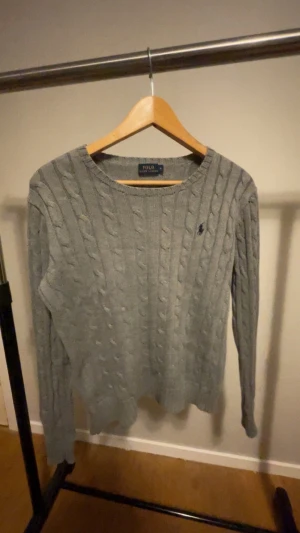 Grå kabelstickad tröja Polo Ralph Lauren - Snygg grå kabelstickad tröja från Polo Ralph Lauren med rund hals och lång ärm. Tröjan har det klassiska broderade logot på bröstet och är gjord i mjuk bomull. Perfekt för dig som gillar stilrena och tidlösa plagg.
