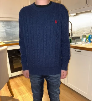  Polo Ralph Lauren - Snygg mörkblå stickad tröja från Polo Ralph Lauren i storlek m. Tröjan har klassiskt flätstickat mönster och en röd broderad logga på bröstet. Rund hals och långa ärmar, perfekt för dig som gillar preppy stil och vill ha något som känns både chill och stilrent.