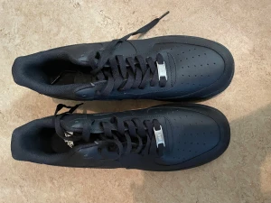 Svarta Nike Air Force 1 sneakers - Säljer ett par svarta Nike Air Force 1 sneakers i klassisk low modell. Skorna har en stilren design med perforerad tå, snörning och platt sula. Tillverkade i skinn med diskret Nike-logga på sidan och metall-detalj vid snörningen. Perfekta för dig som gillar streetstyle.