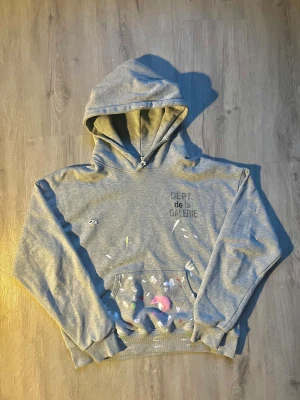 Grå hoodie från Gallery Dept. - Säljer en grå hoodie från Gallery Dept. med tryck på bröstet och coola färgstänk över hela tröjan. Hoodien har huva med snörning, känguruficka och ribbade muddar. Perfekt för dig som gillar streetwear och unika detaljer.