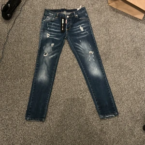 Blå slitna jeansbyxor från Dsquared2 - Säljer ett par blå jeansbyxor från Dsquared2 med slitningar och hål på framsidan. Klassisk femficksmodell med raka ben och knappgylf. Perfekta för en avslappnad och trendig look. Storleken är inte W30 utan storlek 12-13 Years. De är troligen inte f@k3