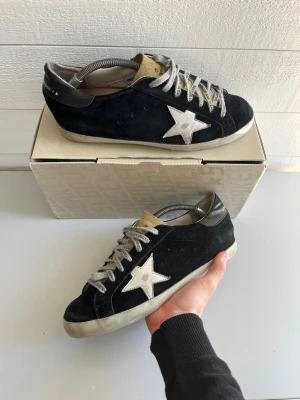 Golden Goose svarta sneakers med stjärna - Golden Goose sneakers i svart mocka med vit stjärndetalj på sidan och beige plös. Skorna har rund tå, platt sula och vita snören med text. Snygg kontrast mellan svart och vitt, perfekt för dig som gillar streetstyle och unika detaljer.