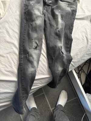 Dsquared2 svarta distressed jeans - Säljer ett par svarta Dsquared2 jeans med slitningar, färgstänk och tvättad look. Jeansen har raka ben, fem fickor och tydlig Dsquared2-logga bak i midjan. Materialet är klassisk denim i bomull och passformen är normal med en modern streetkänsla.