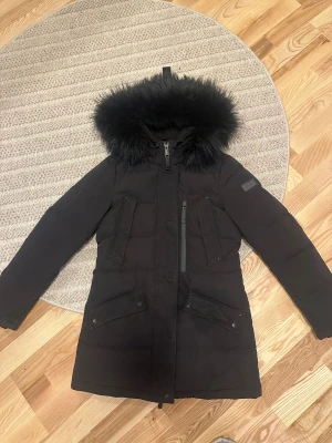 Svart lång dunjacka med päls - Säljer en svart dunjacka med fluffig svart päls på huvan. Jackan är lång, har flera fickor med lock och dragkedja framtill. Perfekt för kalla dagar och har en stilren look som funkar till det mesta. Huvan har avtagbar päls med knappar.