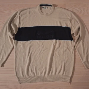 Beige stickad tröja från Lacoste XXL - Snygg beige stickad tröja från Lacoste i storlek XXL. Tröjan har en bred svart rand över bröstet med broderad Lacoste-logga. Rund halsringning med ribbade muddar och klassisk passform. Perfekt för dig som gillar stilren och sportig vibe.