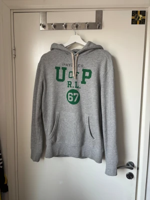 Grå hoodie Polo Ralph Lauren - Grå hoodie från Polo Ralph Lauren med grönt tryck framtill, snörning i huvan och känguruficka. Tillverkad i mjuk bomulls- och polyesterblandning. Klassisk collegekänsla med tryck 'U of P R.L. 67' på bröstet. Perfekt för chill dagar.