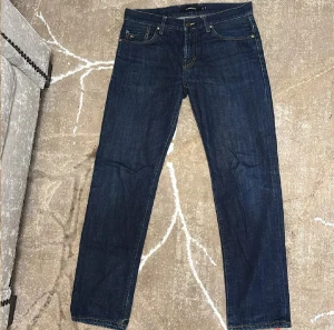 Mörkblå jeans från J.Lindeberg - Snygga mörkblå jeans från J.Lindeberg med klassisk femficksdesign och kontrastsömmar. Jeansen har rak passform och är tillverkade i slitstarkt denim med diskret logga på fickan och läderpatch bak i midjan. Perfekta för en clean och stilren look.