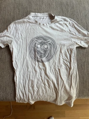 Vit t-shirt från Knowledge Cotton Apparel - Vit t-shirt från Knowledge Cotton Apparel med ett stort svart uggletryck på bröstet. Klassisk rund hals och korta ärmar. Tillverkad i mjuk bomull, perfekt för en clean och stilren look.