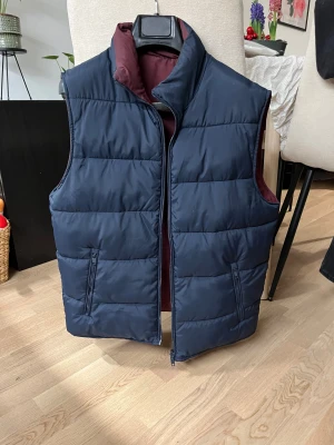 Mörkblå quiltad dunväst - Snygg mörkblå quiltad dunväst med dragkedja framtill och två praktiska fickor med zip. Insidan är vinröd vilket ger en cool kontrast. Perfekt att slänga över en hoodie eller tröja när det är kyligt ute.