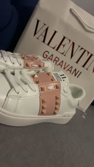 Valentino Rockstud sneakers vit/rosa - Helt nya snygga Valentino Rockstud Untitled sneakers i vitt skinn med ljusrosa band och ikoniska pyramidnitar. Skorna har rund tå, platt sula och vita snören. Perfekt för dig som gillar lyxiga detaljer och vill sticka ut med en unik design.