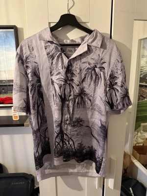 Grå mönstrad skjorta från Zara - Snygg grå skjorta från Zara med svart tropiskt motiv och tunna ränder. Skjortan har korta ärmar, klassisk krage och knäppning framtill. Perfekt för dig som gillar unika prints och vill sticka ut lite extra. Materialet känns lätt och luftigt.