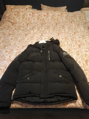 Svart Moose Knuckles dunjacka - Svart dunjacka från Moose Knuckles med pälskant på huvan och ikonisk logga på ärmen. Jackan har flera fickor med dragkedja och tryckknappar, samt quiltad design för extra värme. Perfekt för kalla dagar och har snygga detaljer som metallknappar och robust dragkedja. Endast använd ett par gånger en mycket varm och stilren jacka 