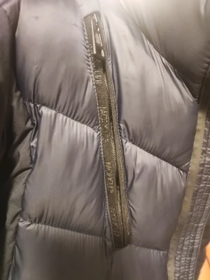 Mörkblå Moncler dunjacka  - Säljer en mörkblå pufferjacka från Moncler med coolt emblem på ärmen och snygga detaljer som dragkedjor på bröstet. Jackan har hög krage och quiltad design, perfekt för kalla dagar. Insidan har en unik serietidningsetikett som ger extra karaktär.