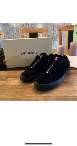 Svarta sneakers Axel Arigato mocka - Snygga svarta sneakers från Axel Arigato i mjuk mocka med dragkedja framtill istället för klassisk snörning. Skorna har en chunky svart platt sula och minimalistisk design som passar till det mesta. Insidan är fodrad med skinn för extra komfort.