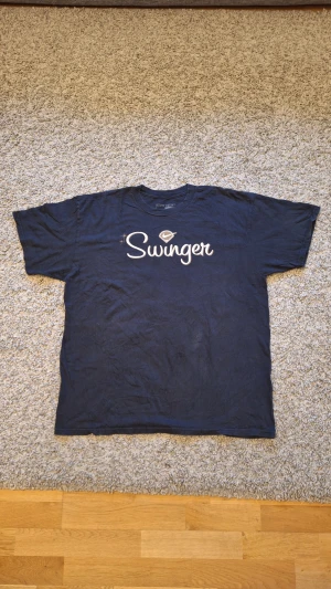 Svart Nike 'Swinger' t-shirt - Snygg svart t-shirt från Nike med "Swinger" text på bröstet i storlek XL, men skulle nog säga den passar mer som L. 100% bomull. Använd fåtal gånger och skicket är jättebra, inga slitningar eller defekter finns. Loggan är i guldig brodyr och skiner. Skick: 8/10