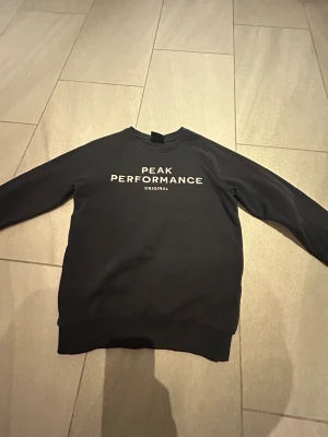 Svart Peak Performance sweatshirt. Storlek 150, pojke - Svart sweatshirt från Peak Performance med vit logga framtill. Klassisk rund halsringning och långa ärmar. Mjuk och skön, perfekt för chill dagar. Ribbad mudd vid ärmslut och nederkant. Enkel och stilren design som funkar till det mesta.