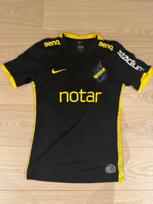AIK svart fotbollströja Nike - AIK:s officiella matchtröja från Nike i svart med gula detaljer och tryck. Kortärmad modell med klubbmärke på bröstet, sponsorloggor och gul kant längs ärmar och sidor. Tillverkad i lätt och ventilerande polyester, perfekt för träning eller match.