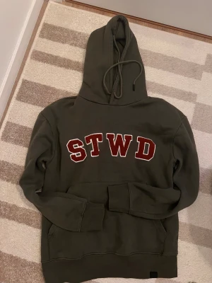 Grön STWD hoodie från Sinsay XS - Säljer en mörkgrön hoodie från Sinsay i storlek XS med STWD-bokstäver i rött och vitt på bröstet. Tröjan har huva med snörning, magficka och långa ärmar. Mjuk insida och klassisk loose passform, perfekt för chill dagar.. passar S också