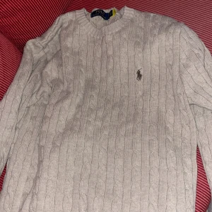 Beige kabelstickad tröja Polo Ralph Lauren - Snygg beige kabelstickad tröja från Polo Ralph Lauren med klassisk rund hals och broderad logga på bröstet. Tröjan har långärmad passform och ribbade muddar vid ärmslut och nederkant. Perfekt för dig som gillar stilrena och tidlösa plagg.