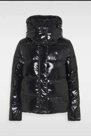 Svart pufferjacka från Calvin Klein - Säljer en svart, glansig pufferjacka från Calvin Klein med hög krage och stor huva. Jackan har CK-logga på bröstet och dragkedja framtill. Den är quiltad och har en loose passform, perfekt för kalla dagar. Snygg och trendig look som funkar till jeans och sneakers.
