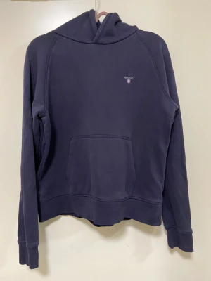 Mörkblå hoodie från GANT - Mörkblå hoodie från GANT med broderad logga på bröstet. Klassisk känguruficka framtill, huva och långa ärmar. Tillverkad i mjuk bomullsmix som är skön att ha på sig. Perfekt för chill dagar eller när du vill ha en enkel och stilren look. Fint skick!