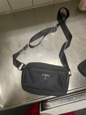 Svart axelväska från Prada med extra pouch - Snygg svart axelväska från Prada i nylon med justerbar rem och silverfärgade detaljer. Väskan har ett klassiskt triangulärt Prada-emblem framtill och en extra liten pouch med dragkedja som kan fästas på remmen. Perfekt storlek för det viktigaste och stilren design. Det ända felet med den är att det gick sunder och jag behövde sticka den på en av ärmena som man ser på bilden
