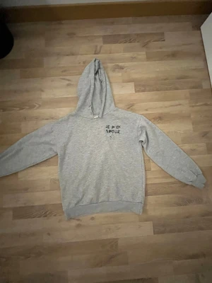 Grå hoodie med tryck från Gina Tricot - Grå hoodie från Gina Tricot med svart texttryck 'JE M'EN AMOUR' och en stjärna på både fram- och baksidan. Tröjan har huva, långa ärmar och ribbade muddar. Mjuk insida i bomullsmix, perfekt för chill dagar. Saknar snören