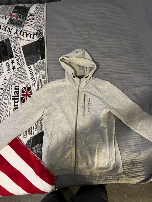 Grå hoodie från Sail Racing - Snygg ljusgrå hoodie från Sail Racing med dragkedja hela vägen och en bröstficka med zip. Tröjan har huva med snörning och ribbade muddar vid ärmslut. Perfekt för chill dagar och enkel att matcha med jeans eller joggers.